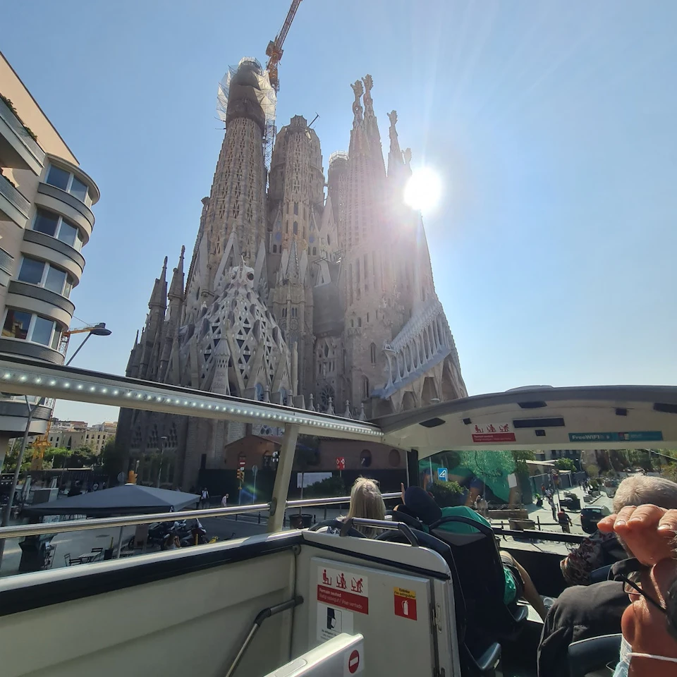 Tourists on an open-top bus viewing the Sagrada Família under clear skies, with a construction crane above the cathedral.