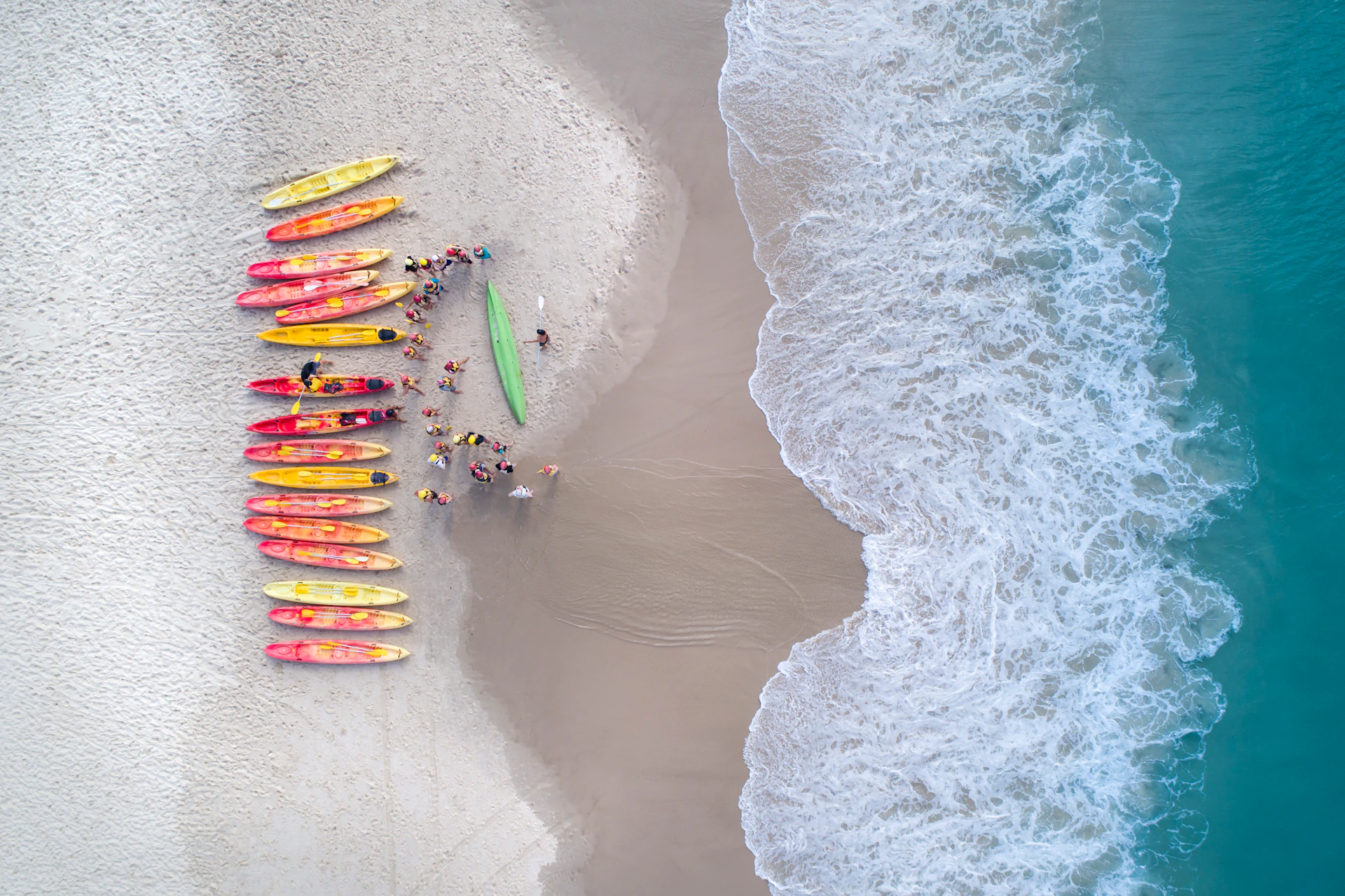 Aerial view of a beach with a row of colorful kayaks and people gathered near the shoreline where waves meet the sand.