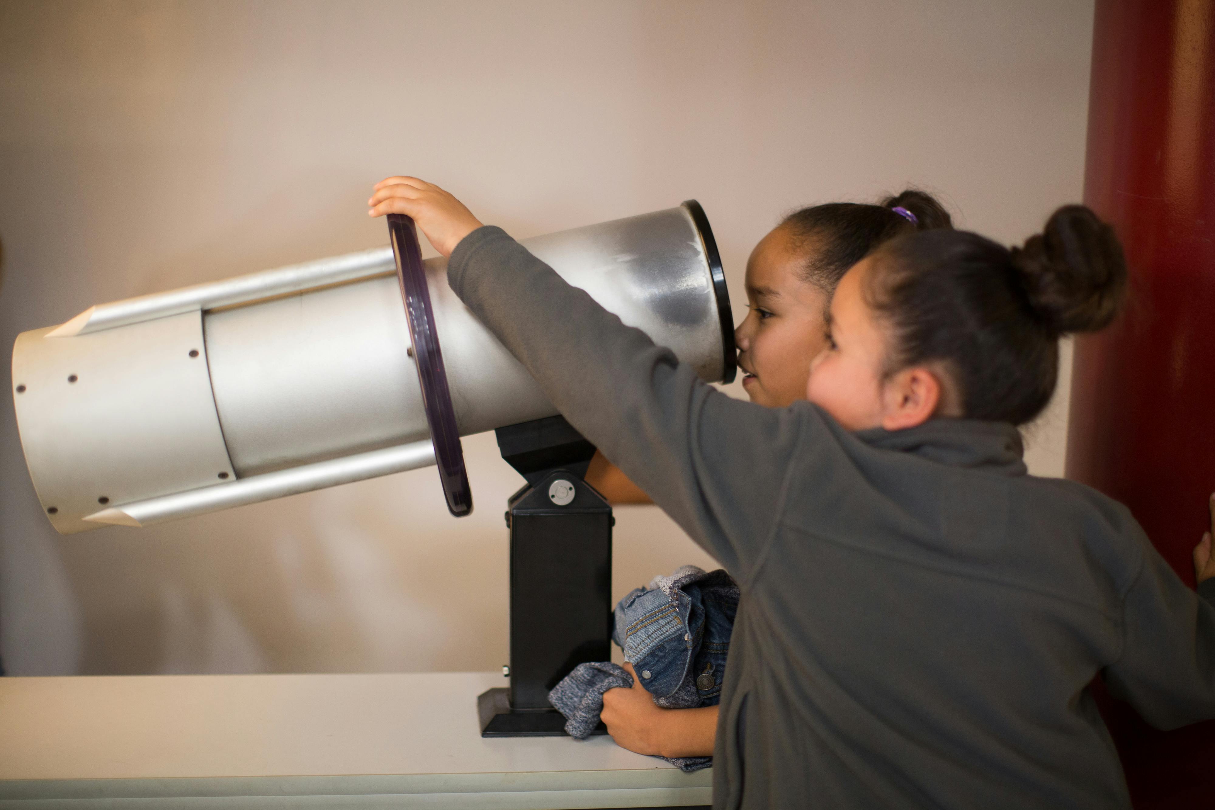 Δύο παιδιά κοιτάζουν μέσα από ένα μεγάλο ασημένιο τηλεσκόπιο μέσα στο σπίτι. Ένα παιδί κρατά ένα μικρό παιχνίδι ή ένα ρούχο.