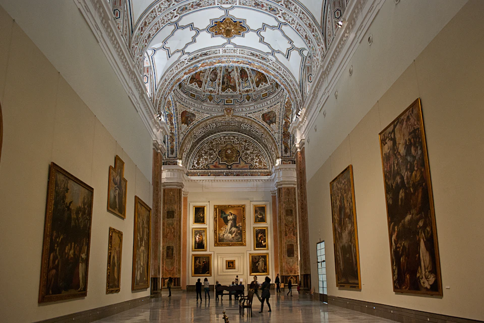 Ornate museum gallery with a decorated ceiling, large framed paintings on walls, and a few visitors observing the artwork.