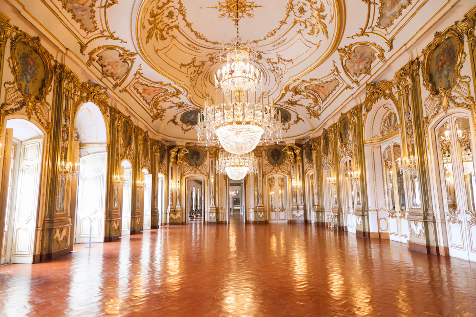 Ornate ballroom with elaborately decorated ceiling, large chandeliers, arched windows, and polished wooden floor.