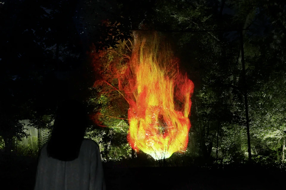 A person observes a large, bright flame projected against a forest background at night.
