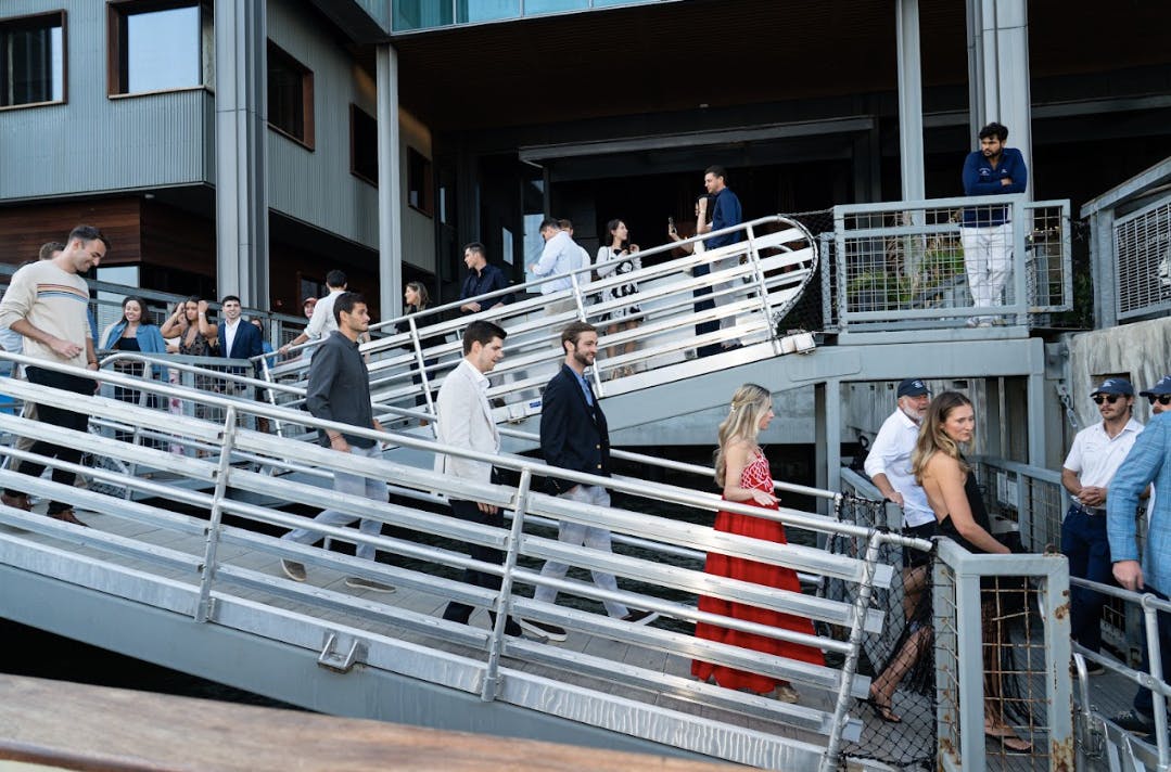 People in formal attire walk on a metal ramp near a building; others stand and converse on upper levels.