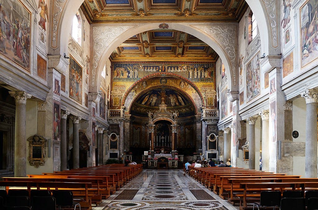 Intérieur d'église orné de fresques peintes, de bancs en bois et de colonnes. Les gens sont assis au fond, près de l'autel.