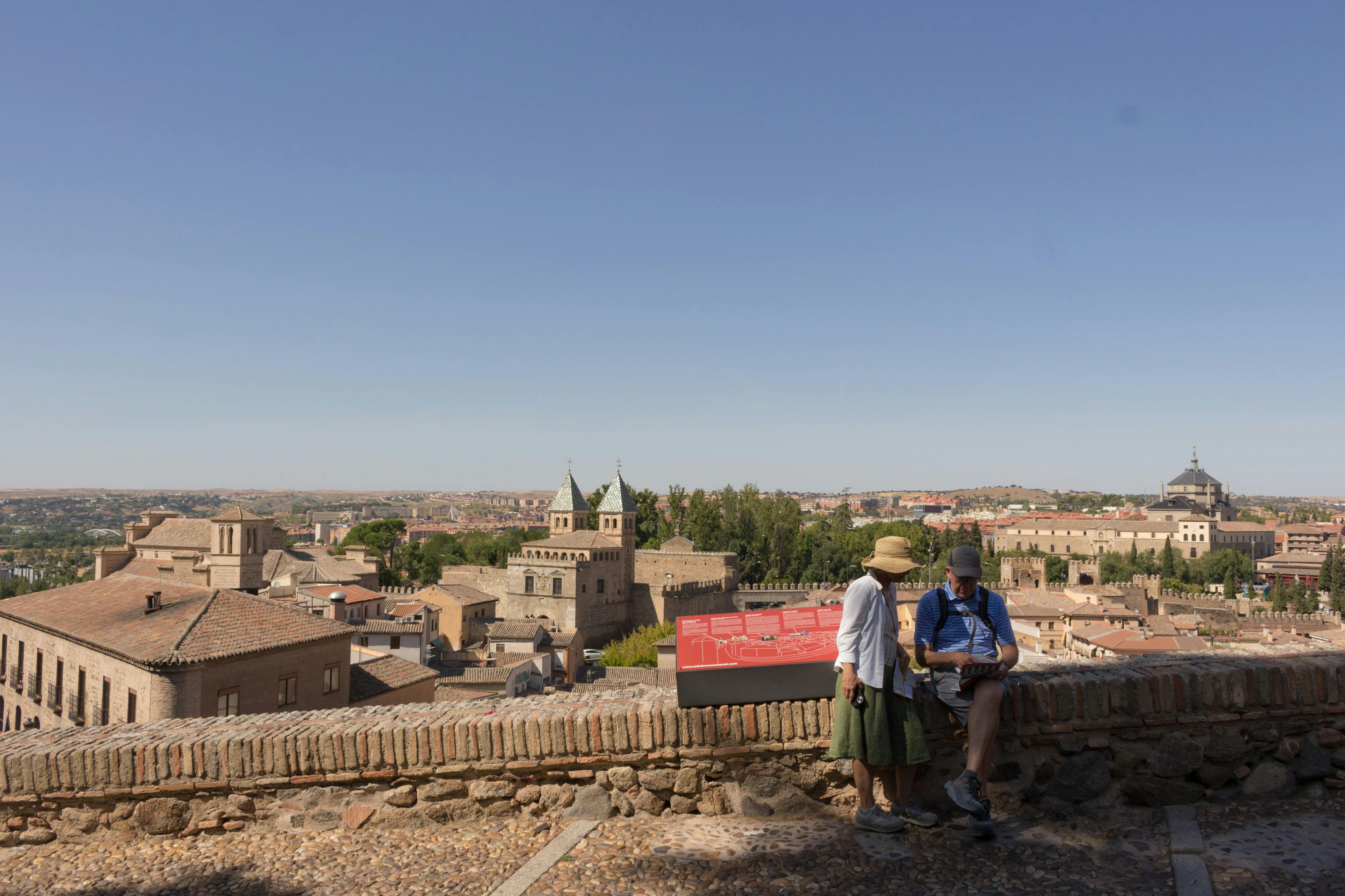 Deux personnes debout près d'un mur de pierre surplombant un paysage urbain historique avec une église et d'autres bâtiments sous un ciel dégagé.