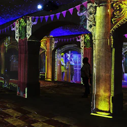 Svagt upplyst rum med välvda stenpelare, färgglada projektioner på väggar och några personer som observerar den uppslukande ljusdisplayen.