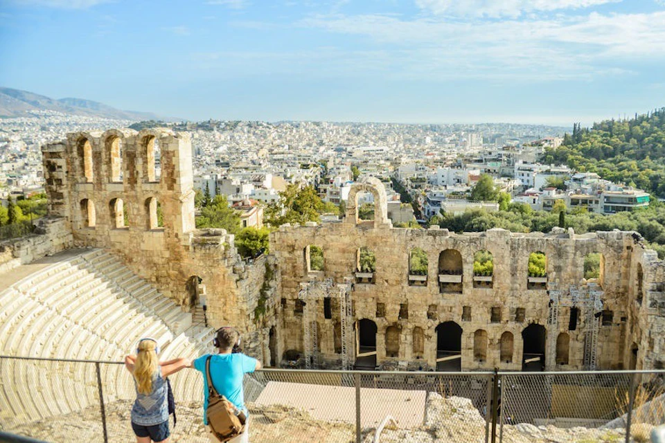 Two people observing an ancient stone amphitheater with a cityscape in the background on a sunny day.