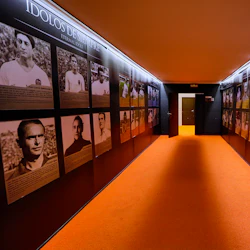 En hall med orange matta, med väggar som visar porträtt och information om fotbollslegender. En delvis öppen dörr är i slutet.