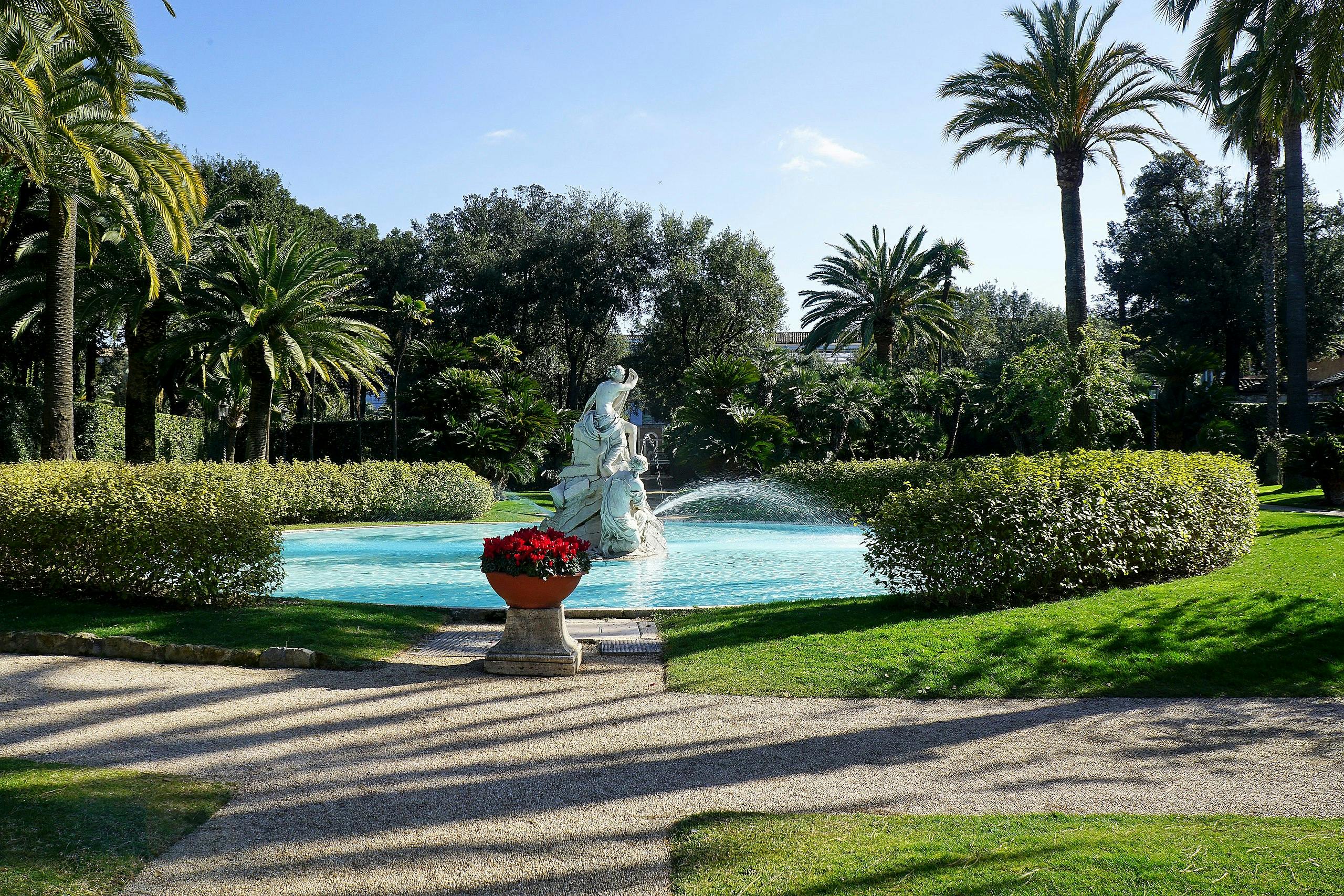 A statue in a garden fountain surrounded by lush greenery, palm trees, hedges, a gravel path, and a potted plant with red flowers.