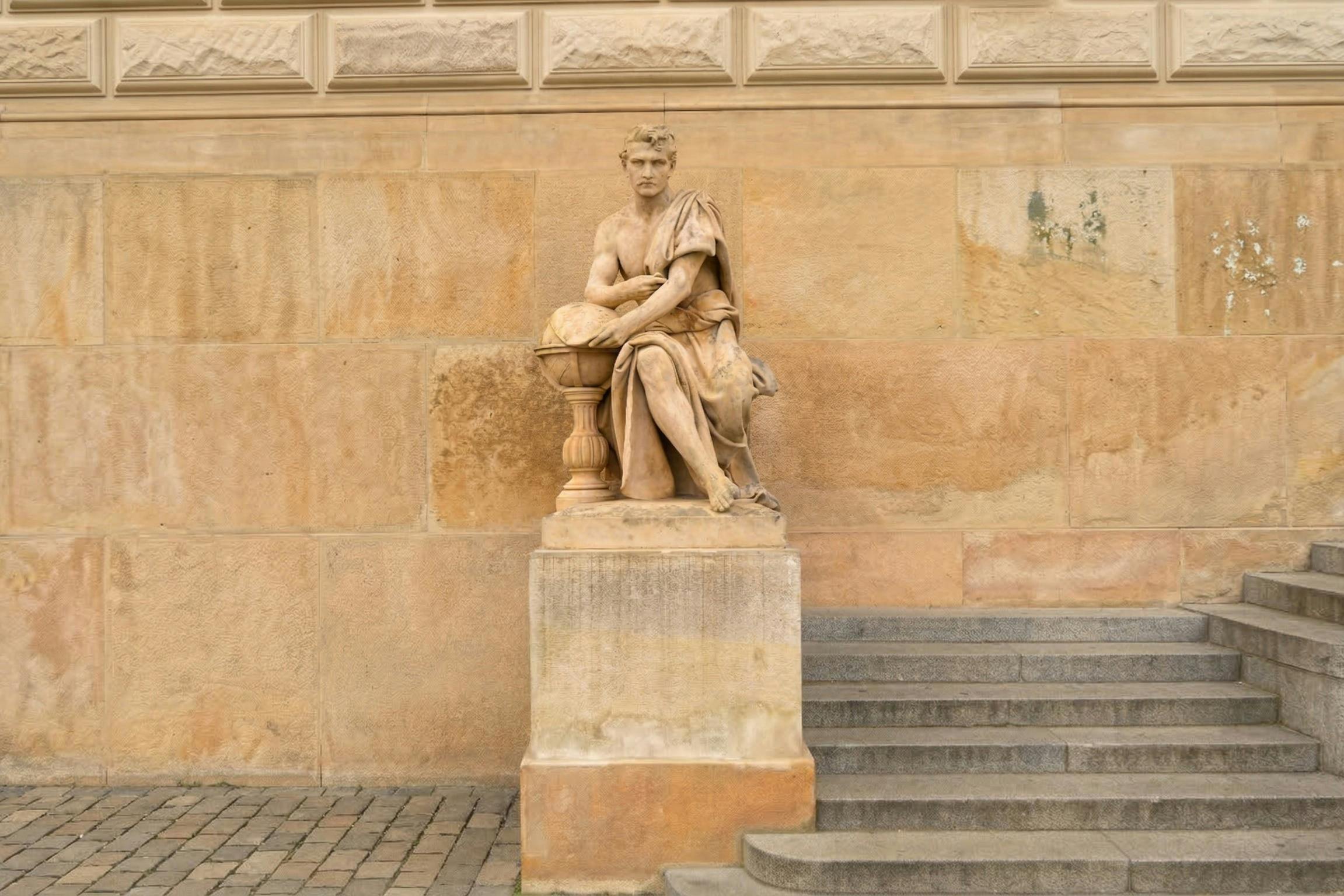 가까이에 계단이 있는 베이지색 벽에 대고 받침대에 놓인, 지구본을 들고 있는 앉은 모습의 석상.