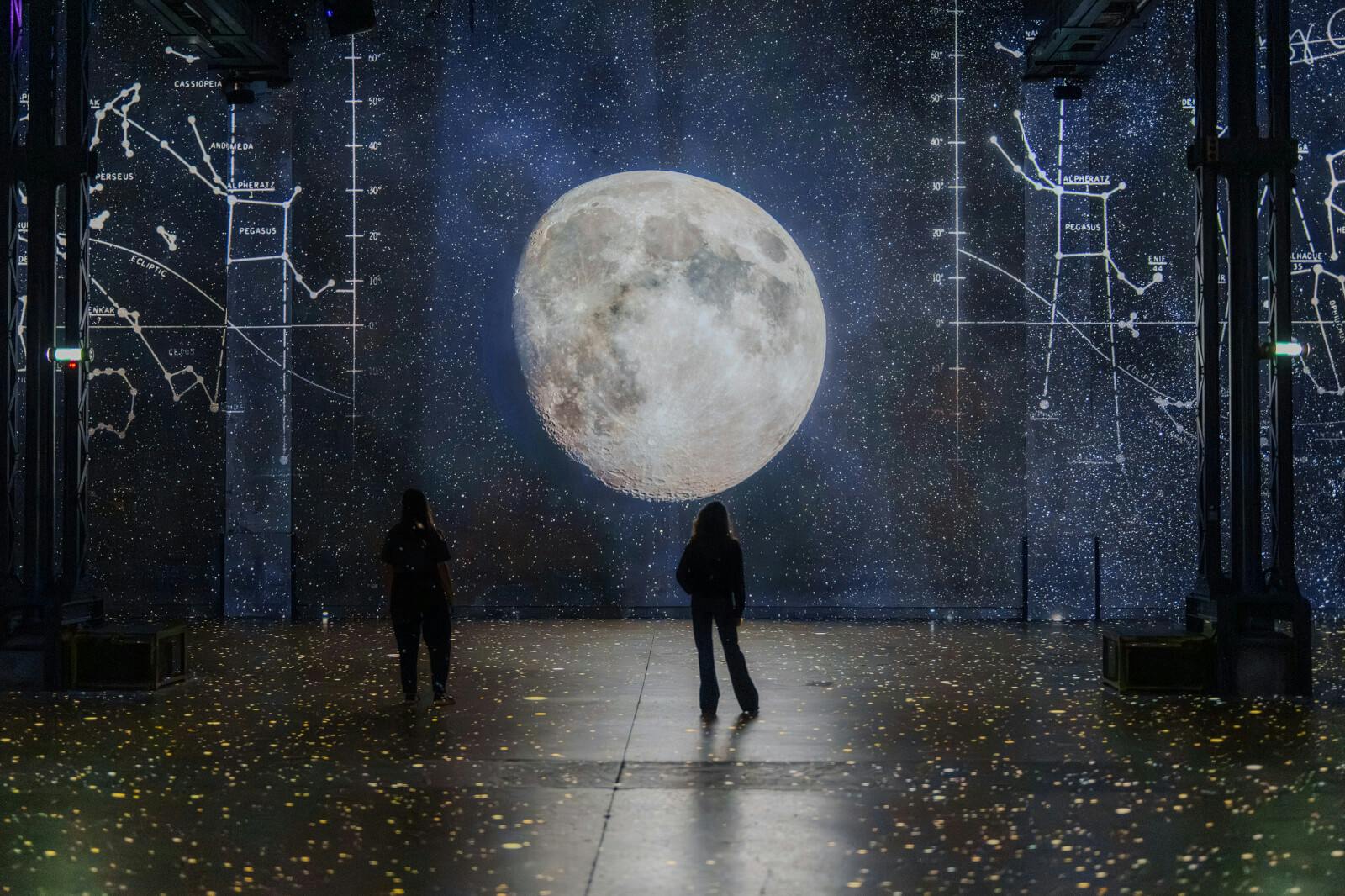 Two people stand on a starry floor, facing a large, detailed moon projection with star charts in the background.
