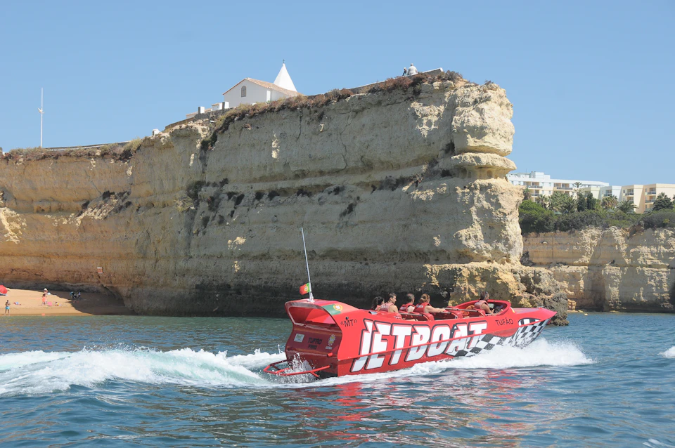 A red jet boat with passengers speeds past a rocky cliff with a building on top, under a clear blue sky.