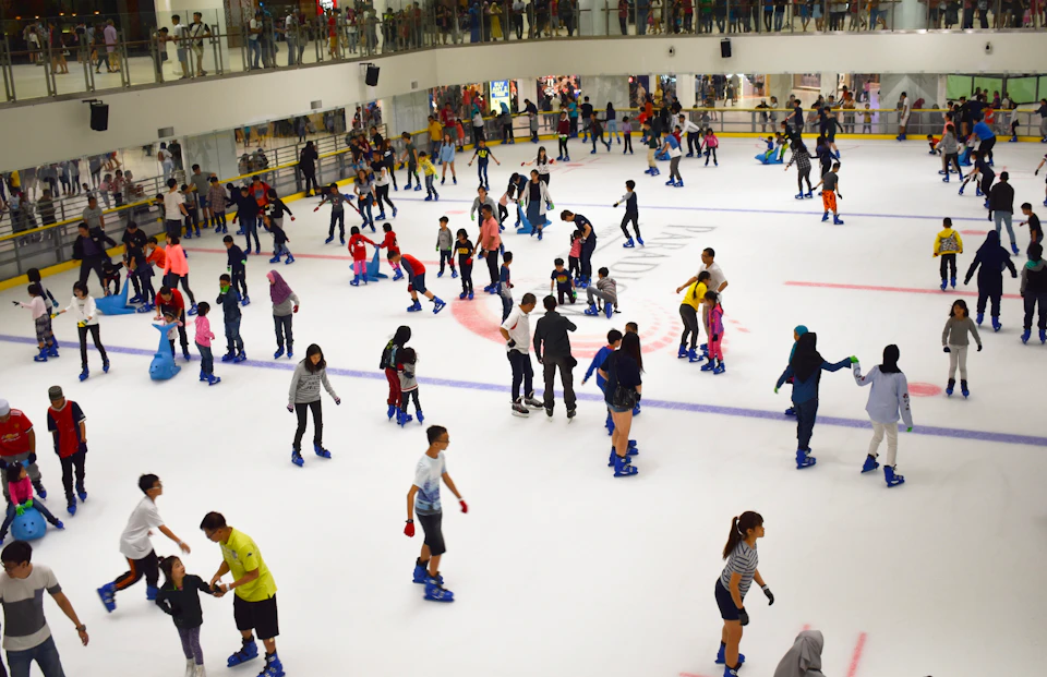 A busy indoor ice skating rink with many people skating and a second-floor balcony with spectators.