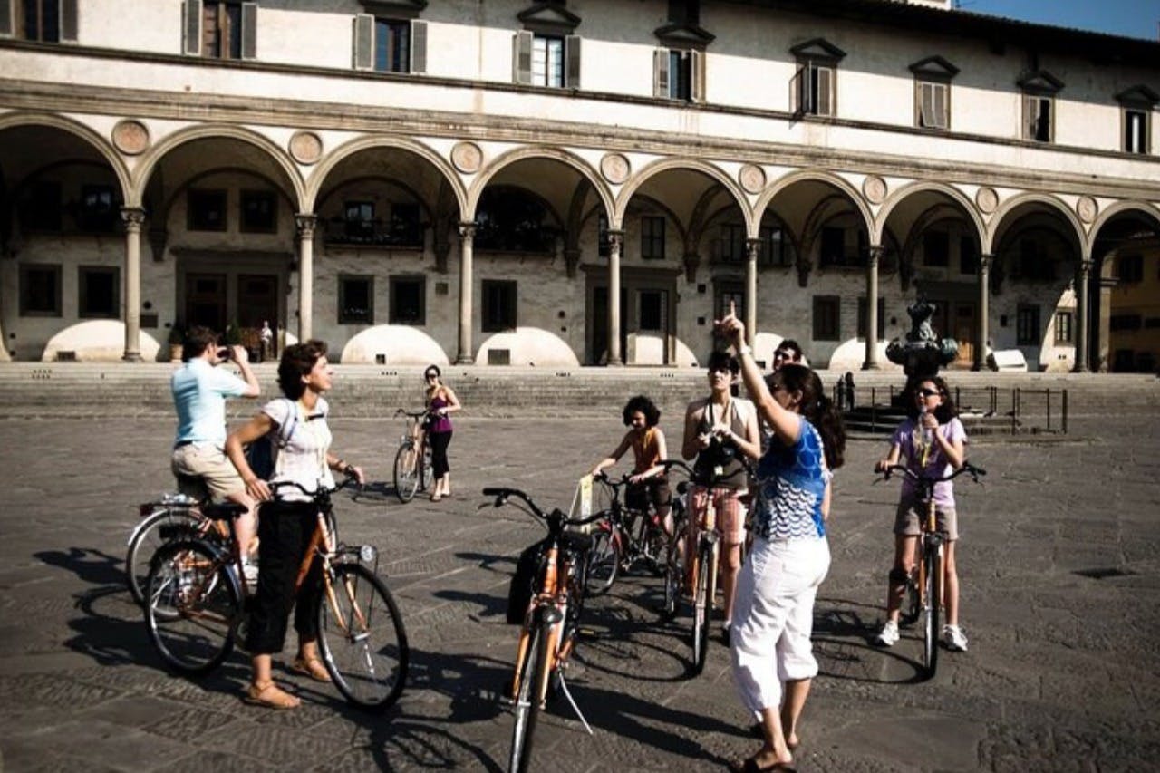 A group of people with bicycles gathered in front of an old building with arched columns.
