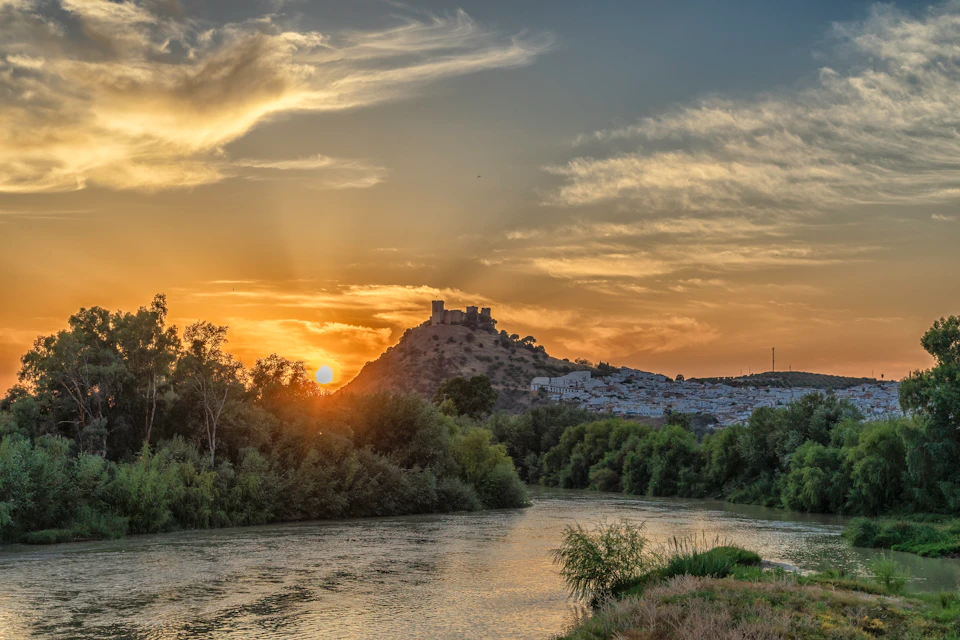 Sunset over a hilltop castle with a nearby river, surrounded by lush trees and a partly cloudy sky.