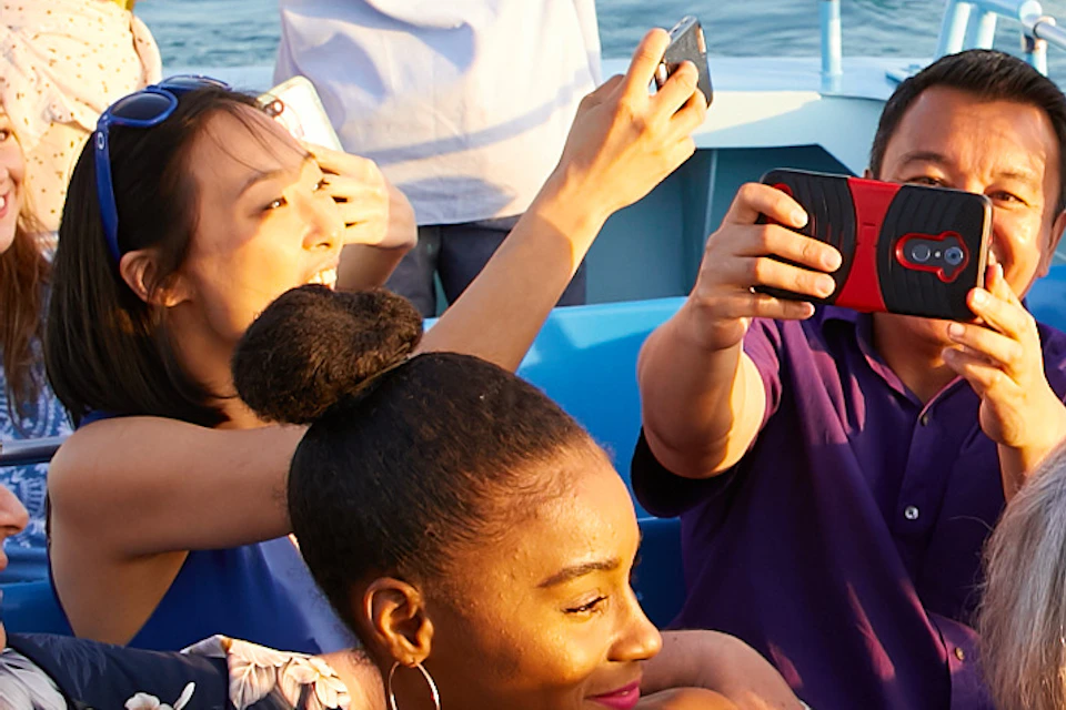 People on a boat take photos with a city skyline in the background. An American flag waves at the stern. The weather is sunny.