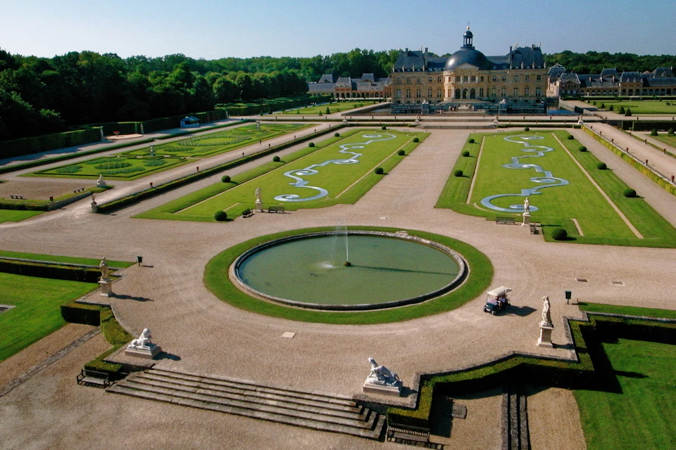 A formal garden with symmetrical lawns, decorative patterns, a central fountain, statues, and a large chateau in the background.