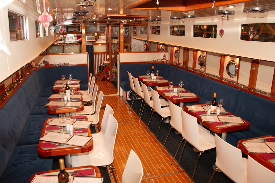 A cozy dining area inside a boat with wooden floors, white chairs, and tables set with menus, glasses, and wine bottles.