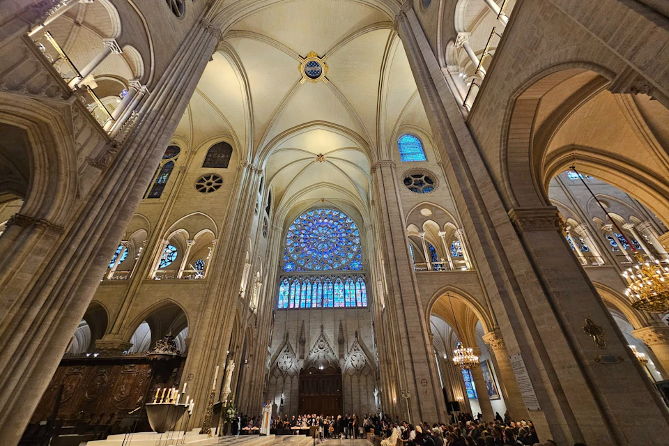 Interior of a large cathedral with high vaulted ceilings, stained glass windows, arched doorways, and a crowd of people gathered below.