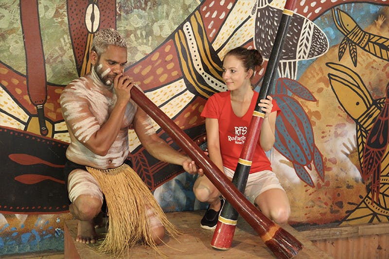 A man plays a didgeridoo while a woman kneels beside him holding another didgeridoo, with a colorful mural in the background.