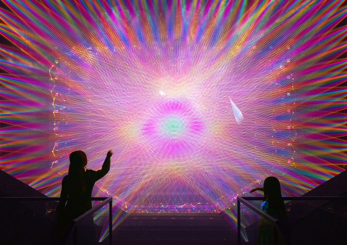 Dues figures de silueta interactuen amb una pantalla de llum vibrant i multicolor que projecta patrons concèntrics.