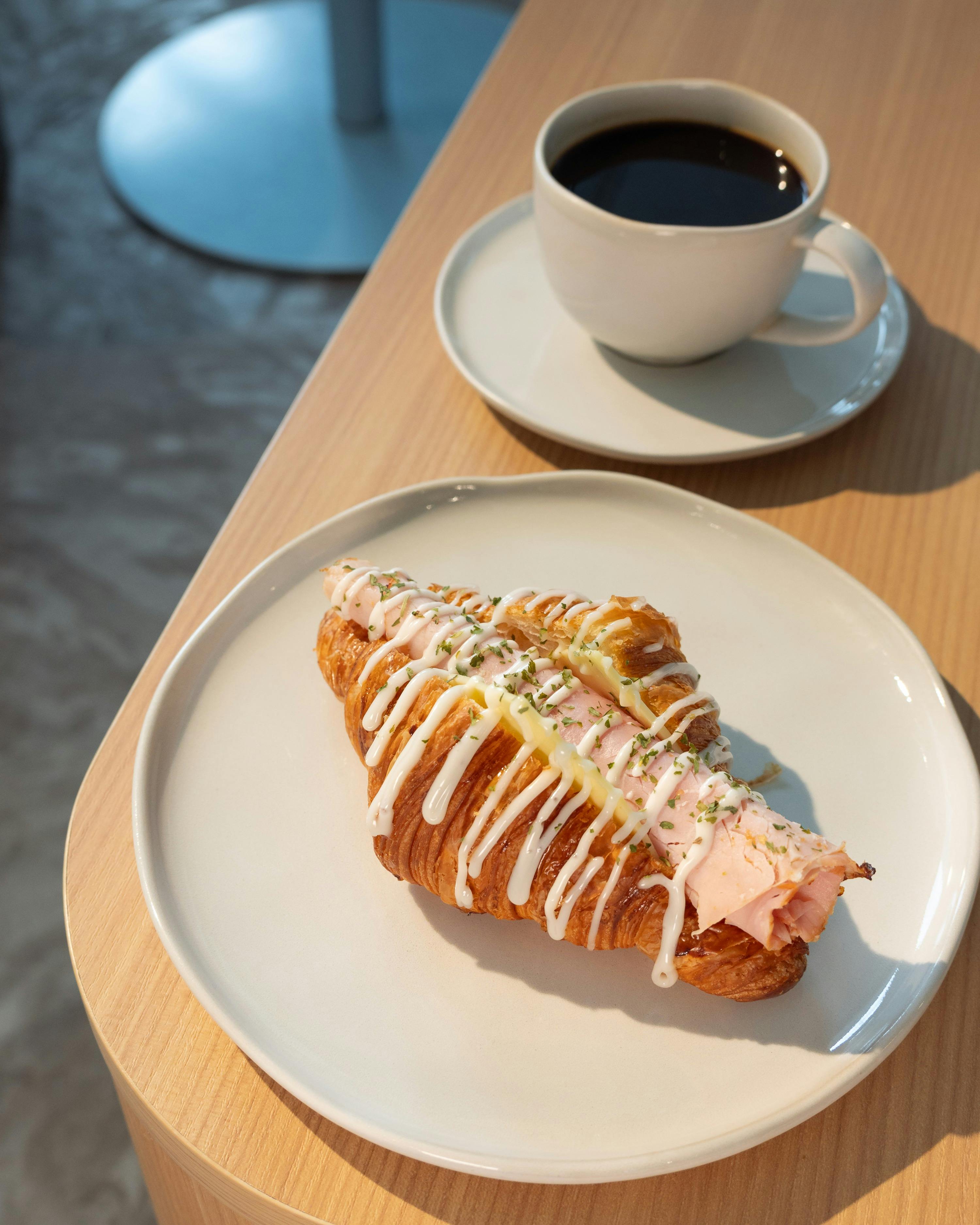 A croissant with pink filling and white drizzle on a plate, next to a cup of black coffee on a wooden table.