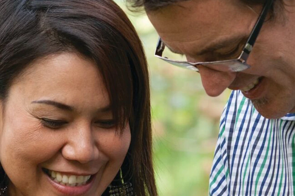 Two people in a kiwi orchard, one holding a cut kiwi fruit, surrounded by hanging kiwi vines.