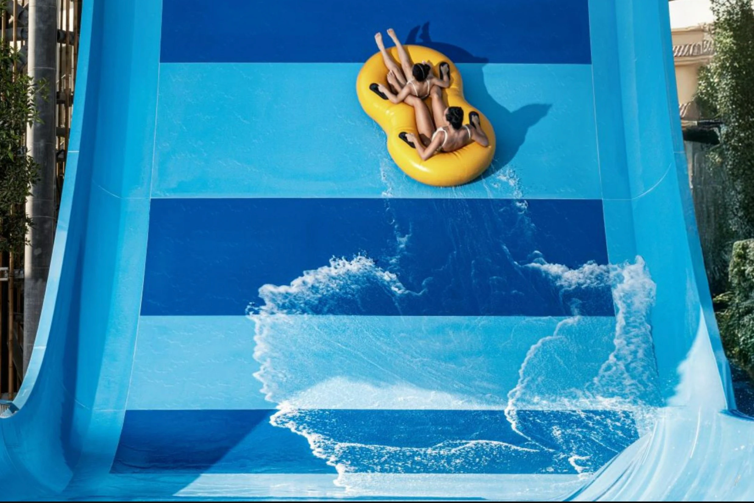 Two people on a yellow inflatable raft sliding down a blue water slide with splashes of water.