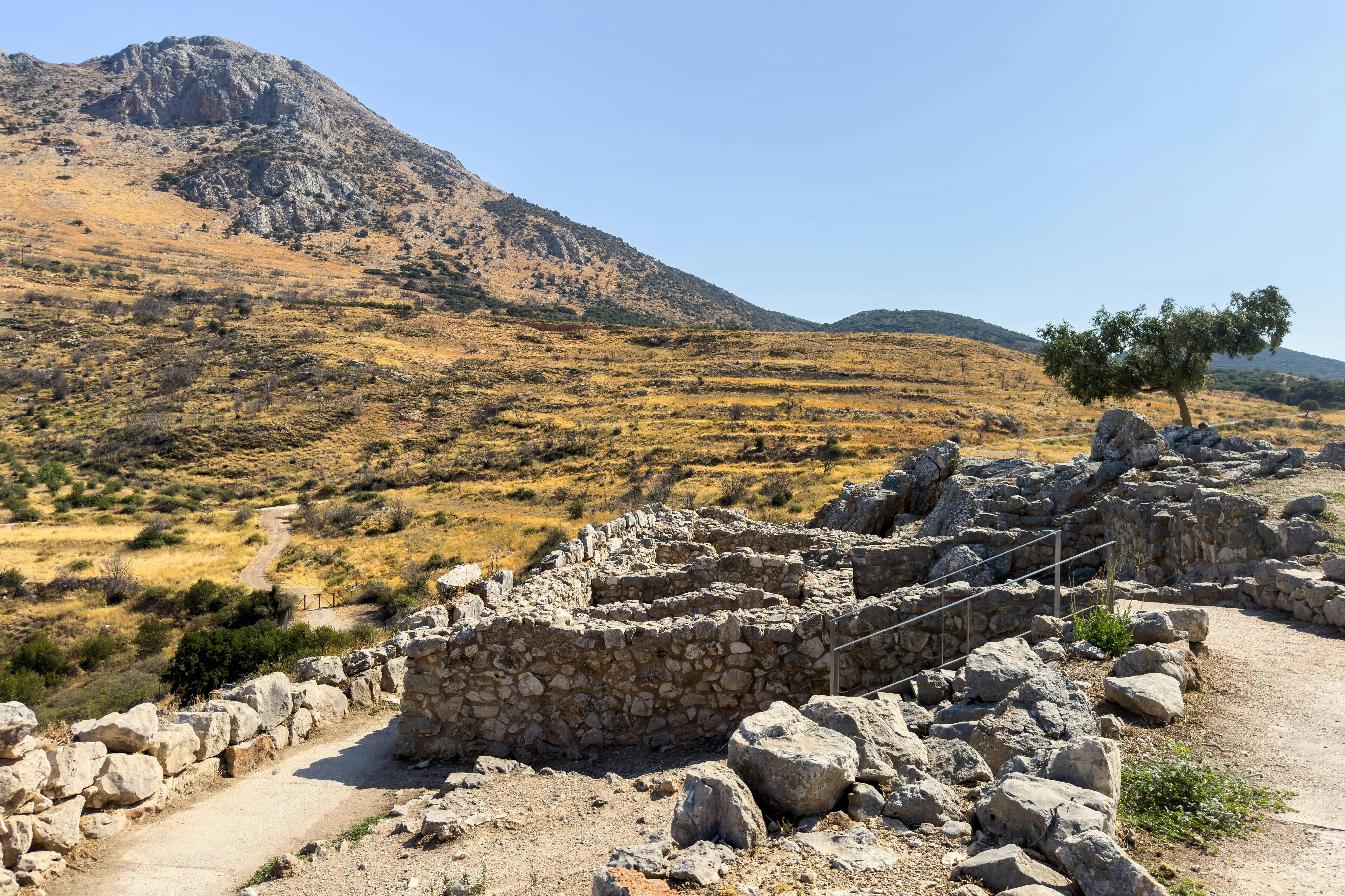 맑고 푸른 하늘 아래 건조하고 언덕이 많은 풍경의 석조 유적지로, 통로와 희박한 초목이 보입니다.
