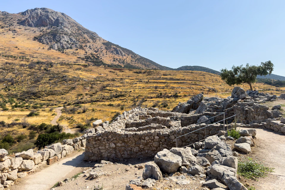 A stone ruin in a dry, hilly landscape under a clear blue sky, with pathways and sparse vegetation visible.