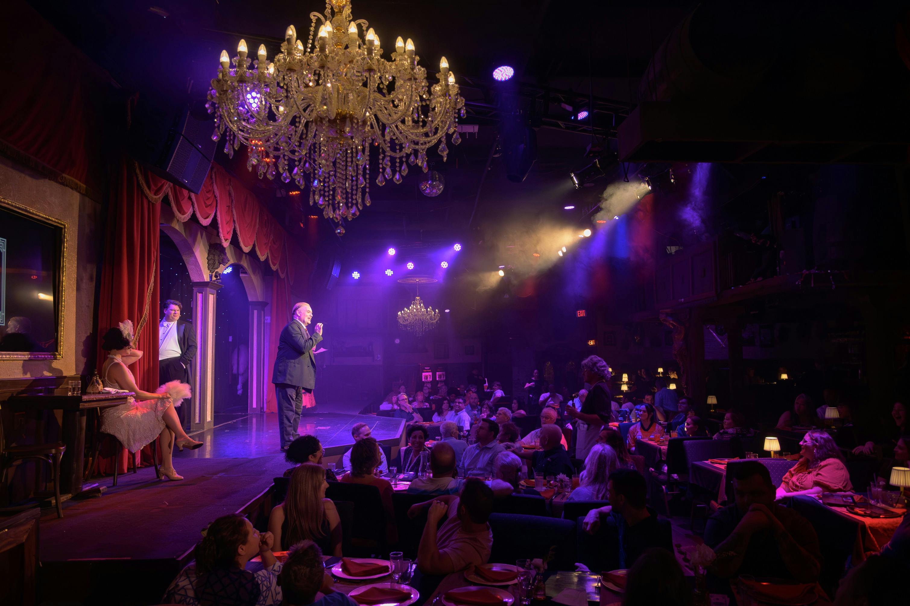 Un home parla en un escenari a un públic en un lloc poc il·luminat amb candelabros i il·luminació escènica de colors.
