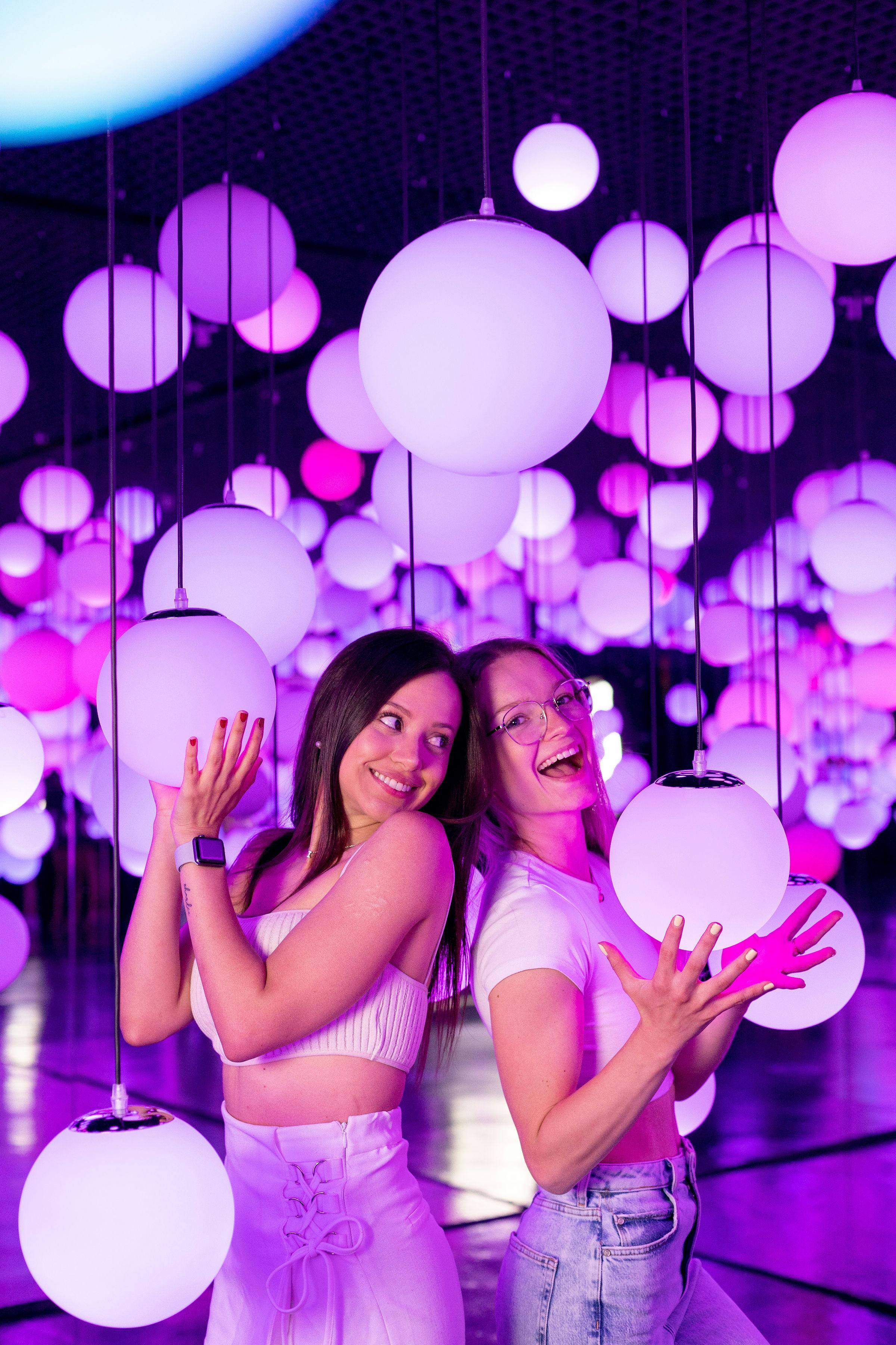 Two women surrounded by large, glowing, pink and purple orbs smile and pose, creating a vibrant and playful atmosphere.