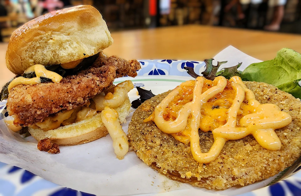 A sandwich with fried chicken and macaroni next to a fried green tomato topped with sauce on a plate with greens.