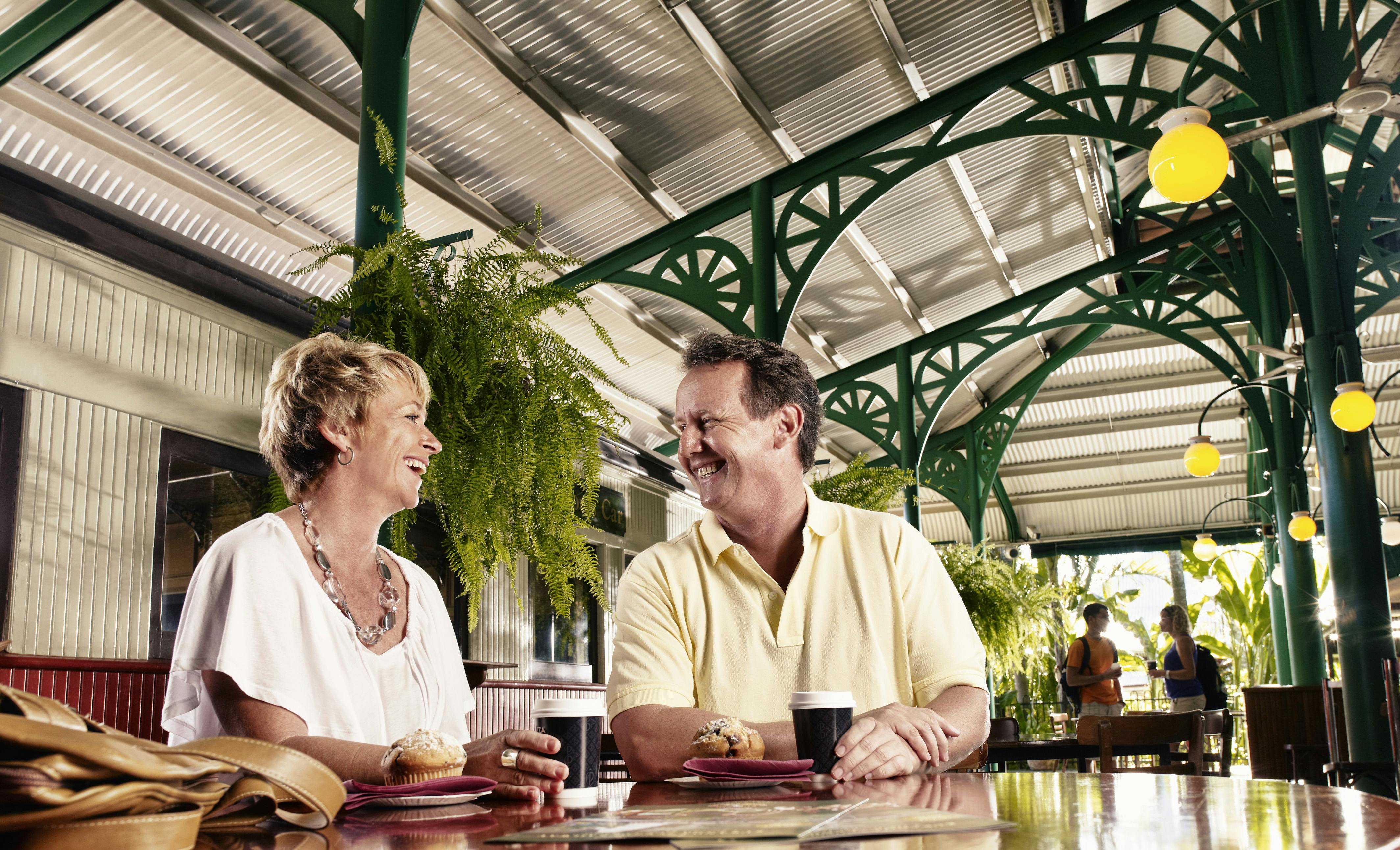 Two people sitting at a table with pastries, smiling at each other, under a metal and green-beamed roof with hanging ferns.