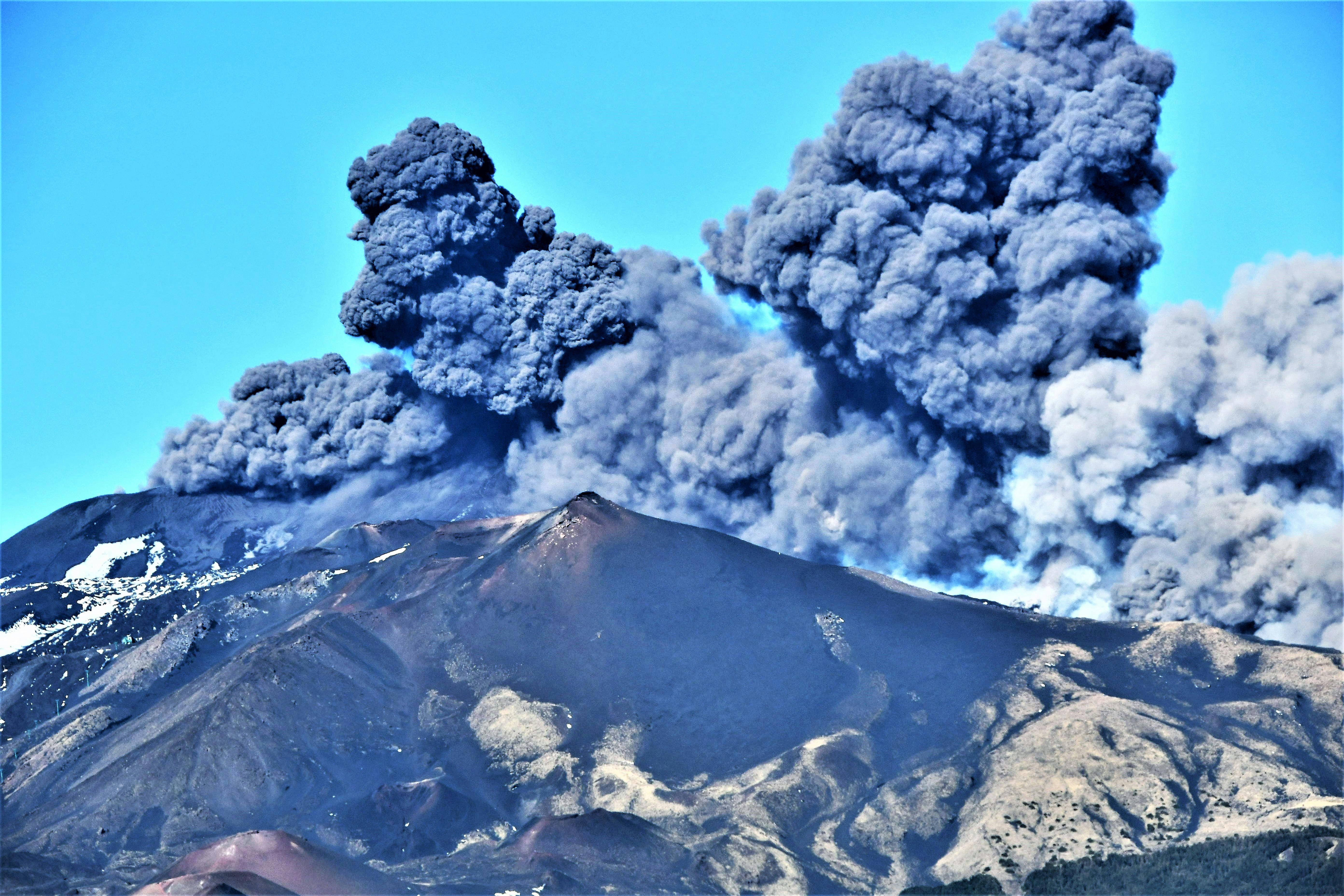 Dense e scure nuvole di fumo e cenere si levano da un vulcano in eruzione sotto un cielo limpido e azzurro.