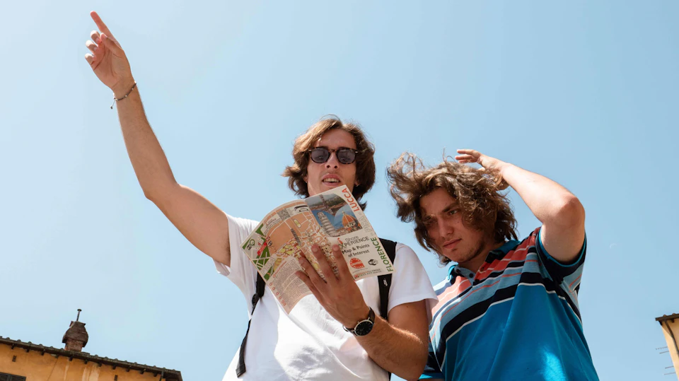Two people outdoors, one wearing sunglasses and holding a map, both looking around under a clear blue sky.