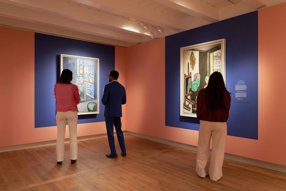 Three people viewing paintings in a gallery with pink and blue walls and wooden flooring.