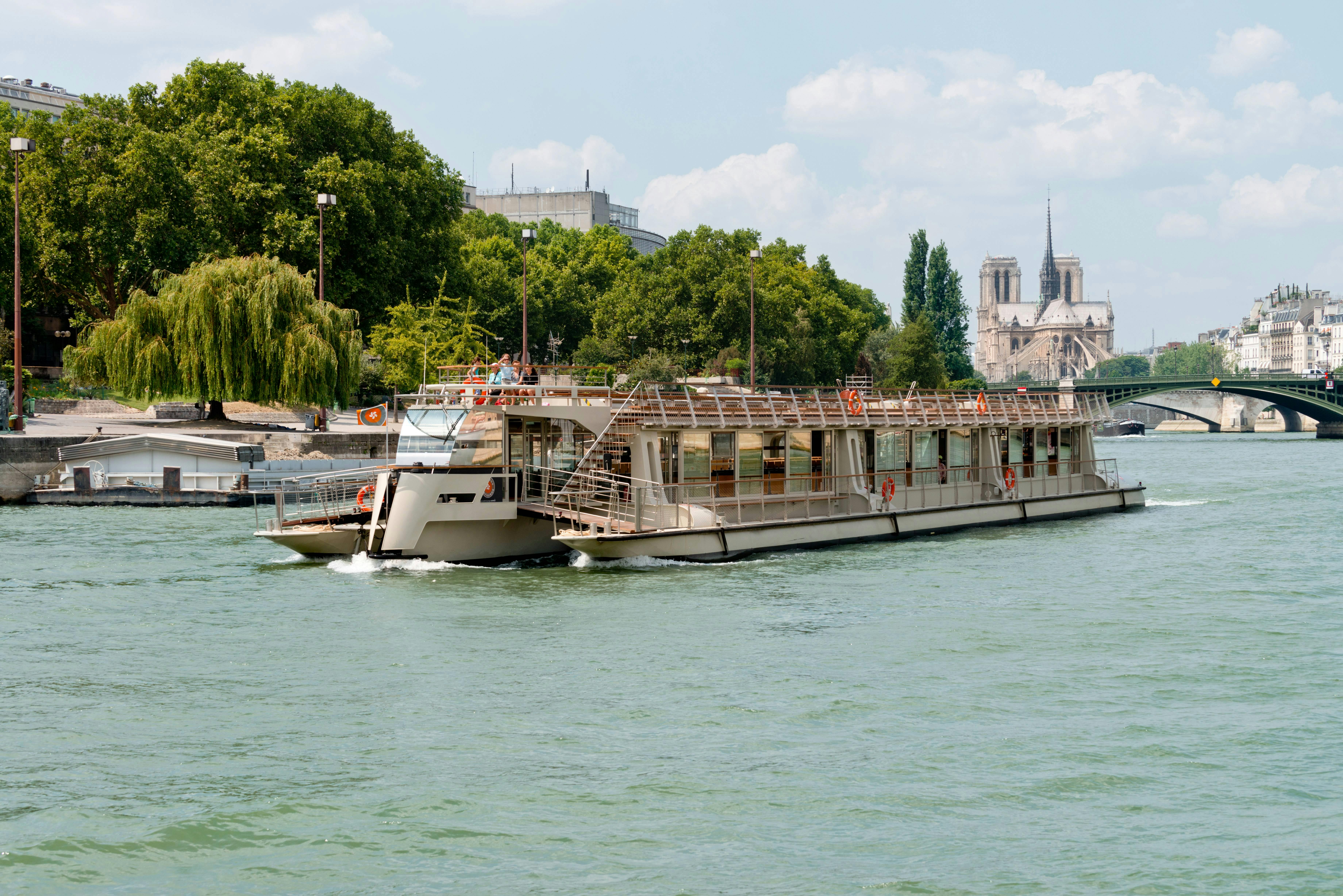 A riverboat cruising on a river with trees in the background, and a cathedral visible in the distance under a clear sky.