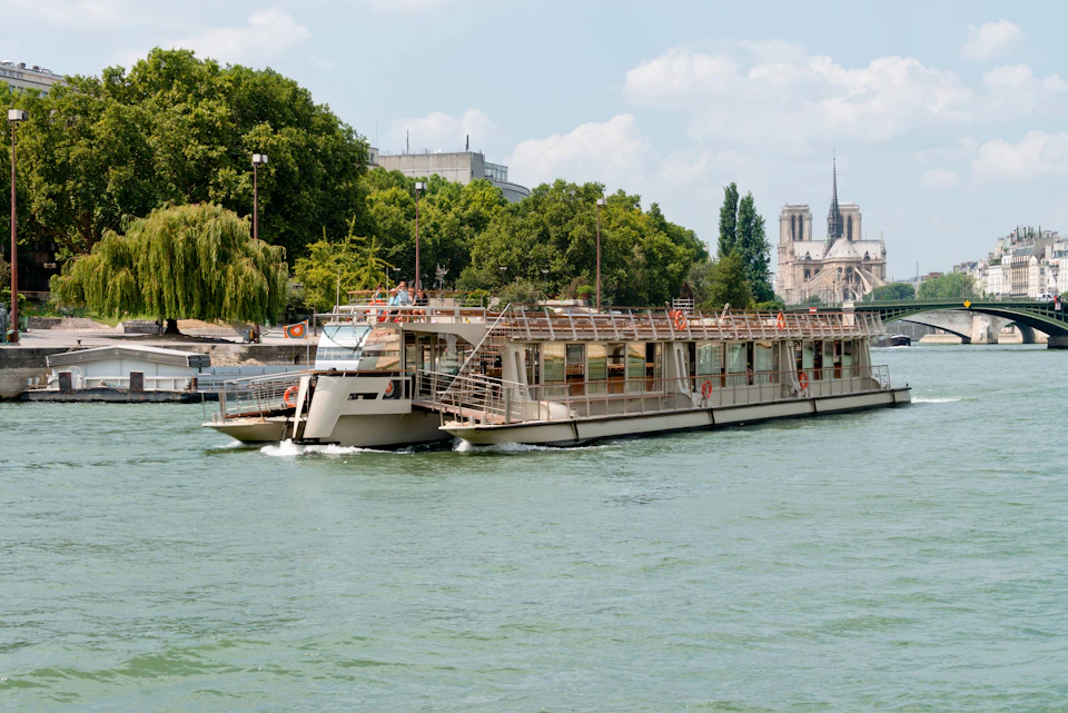 A riverboat cruising on a river with trees in the background, and a cathedral visible in the distance under a clear sky.