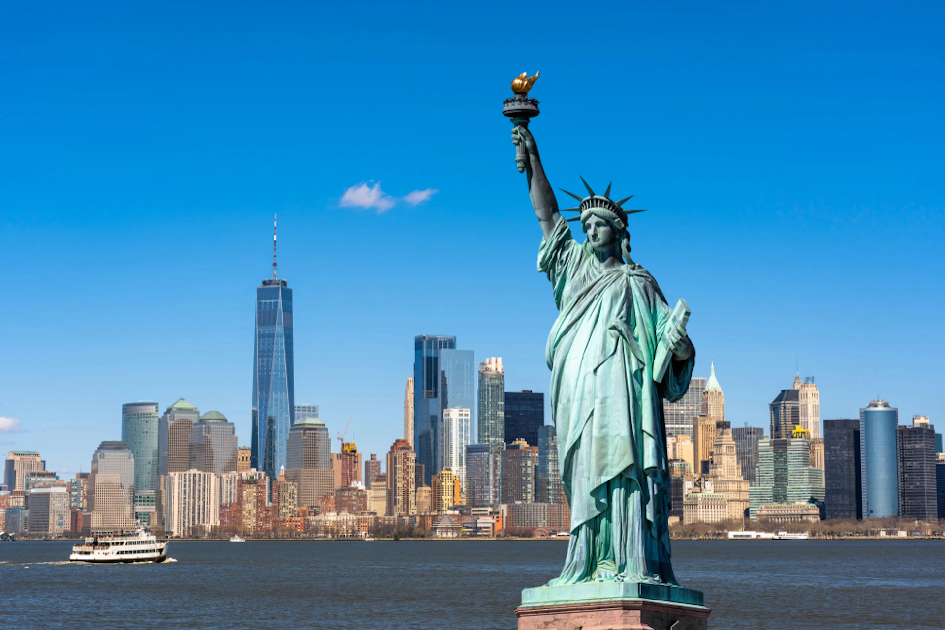 The Statue of Liberty with New York City skyscrapers, including One World Trade Center, in the background under a clear blue sky.