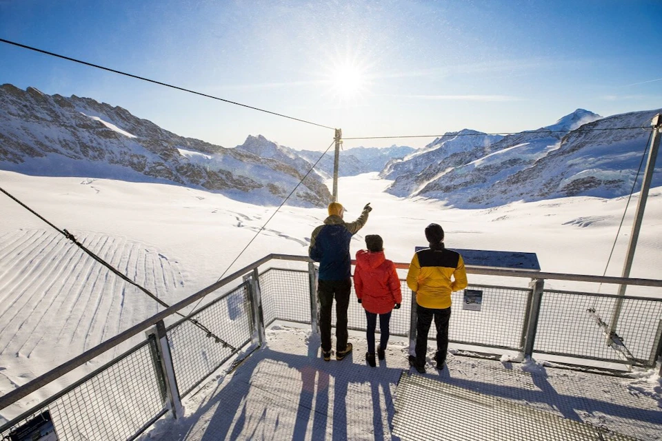 Three people standing on a metal platform overlooking a snowy mountain landscape under a bright sun.
