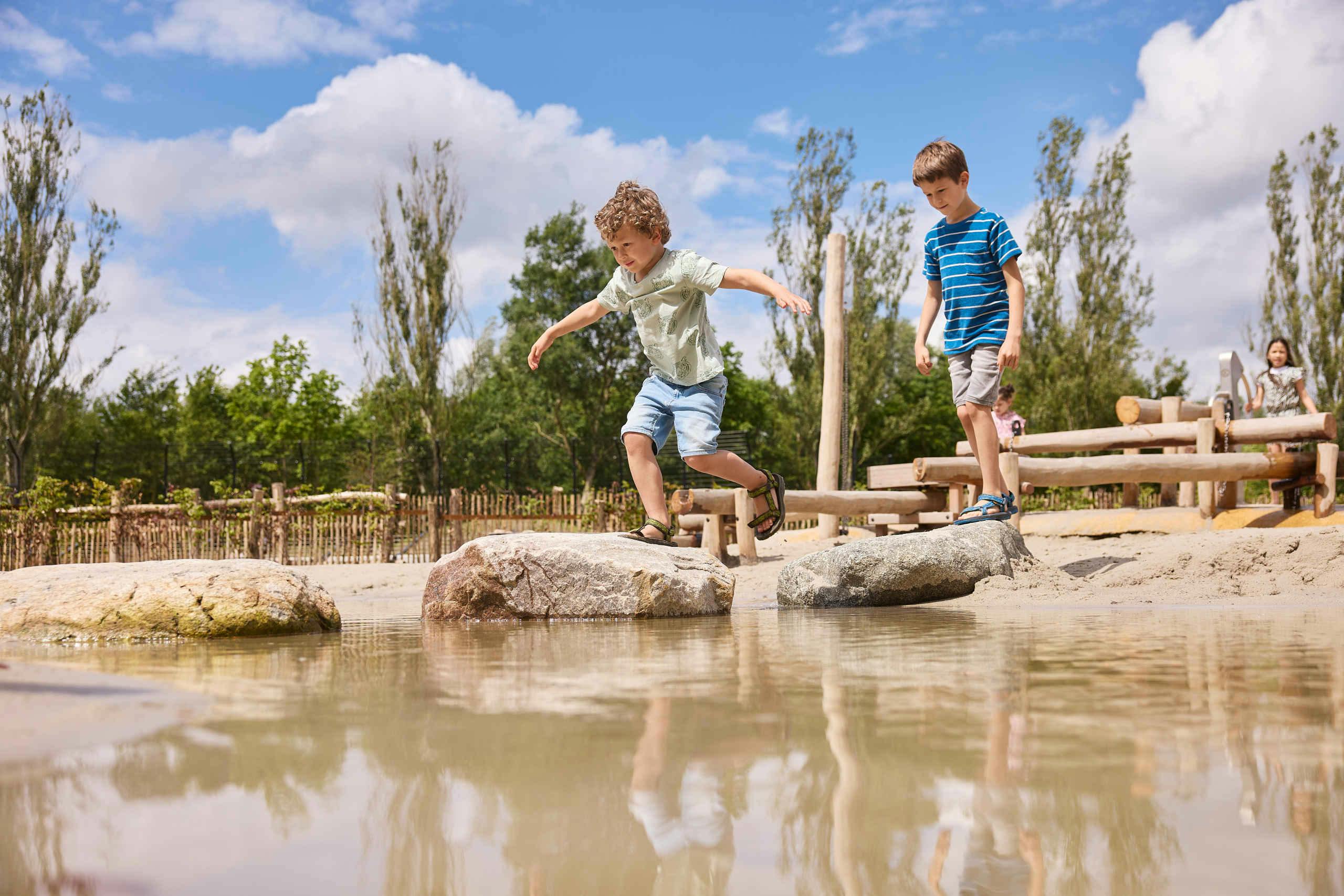Two boys play by a shallow pond, one jumping between rocks while the other watches. Trees and a wooden fence are in the background.