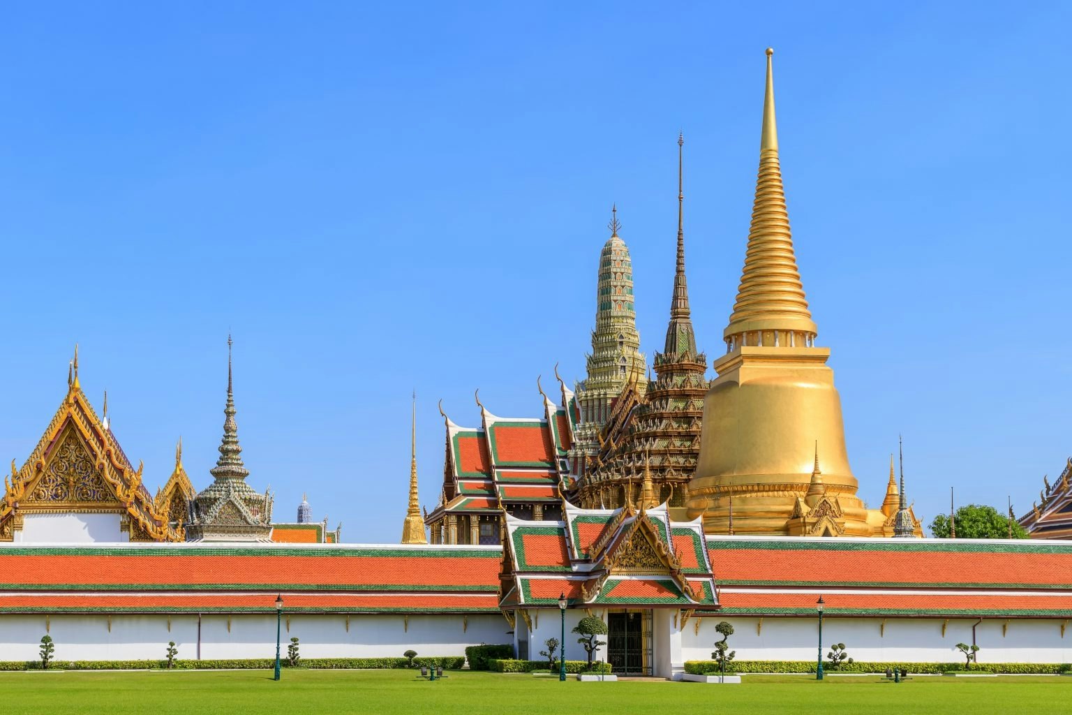 Pagode dorée et temples ornés et colorés sous un ciel bleu clair, entourés d'une pelouse manucurée et d'un mur blanc.