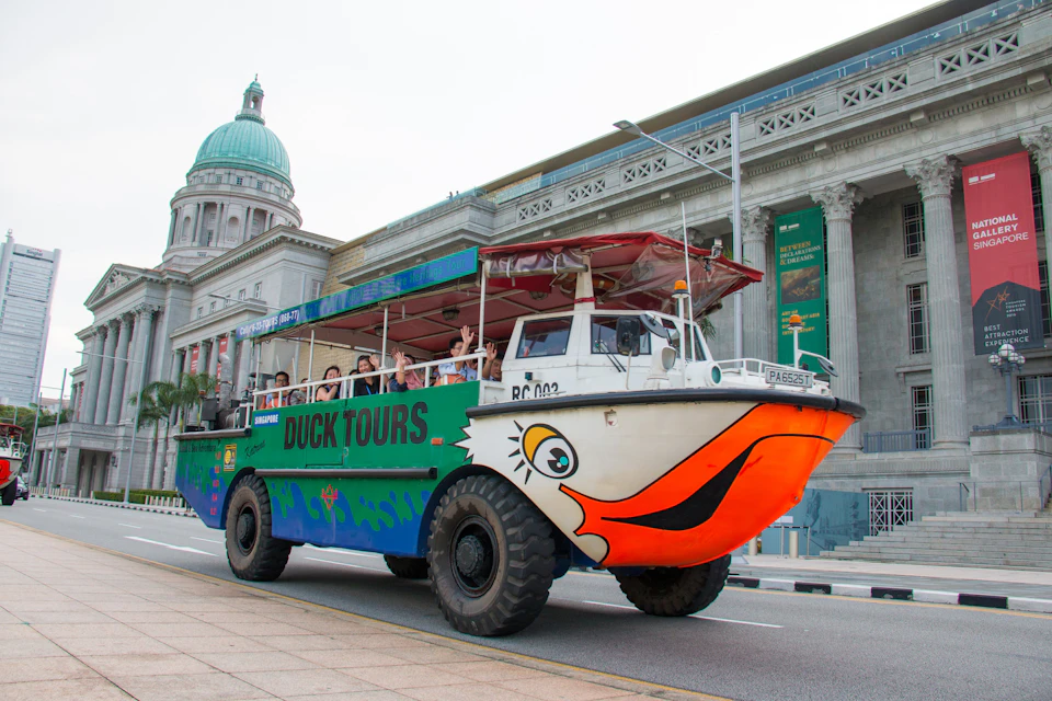 A colorful amphibious tour vehicle labeled "Duck Tours" drives near a historic building with a green dome. Tourists are visible inside.