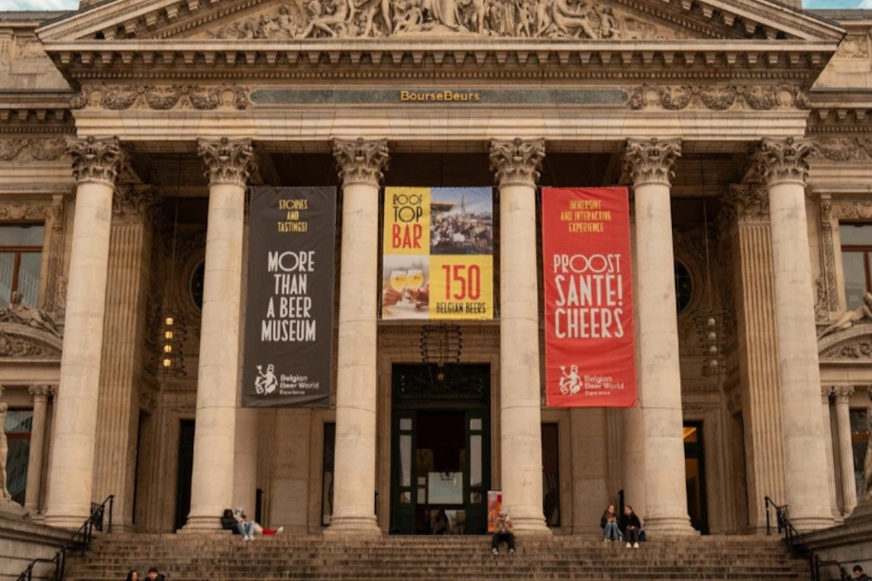 Facade of a historical building with columns, banners promoting a beer museum, and people sitting on the steps.