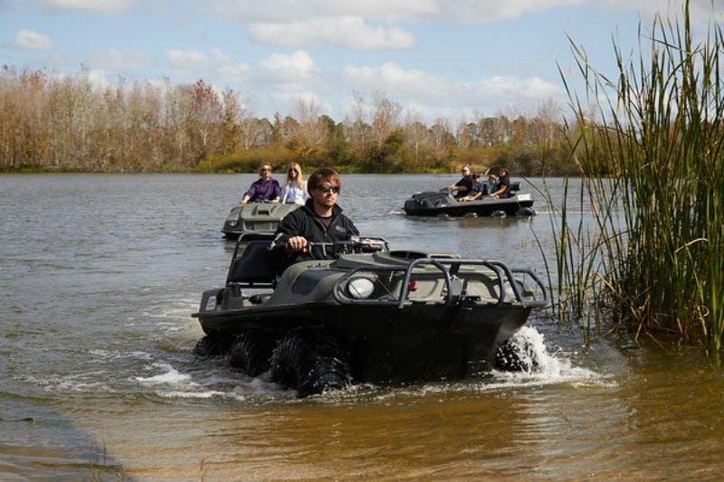 People driving amphibious ATVs on a lake with a forested shoreline in the background, under a partly cloudy sky.