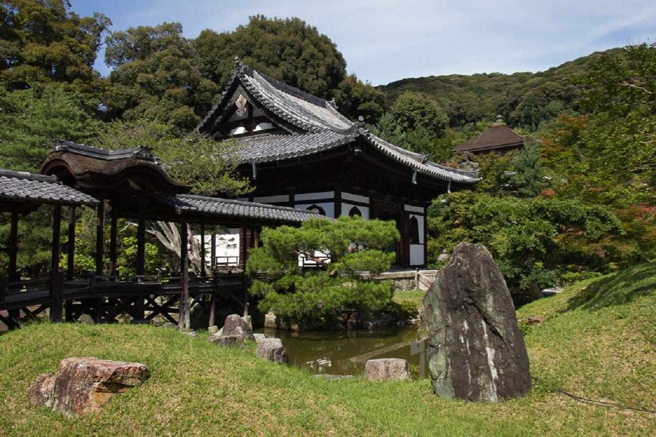 Bâtiment japonais traditionnel avec un toit en tuiles entouré d'une végétation luxuriante, de rochers, d'un petit étang et d'un pont en bois.