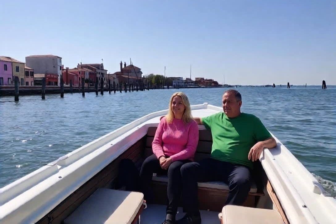 Due persone sedute su una barca in uno specchio d'acqua calmo, con edifici e alberi visibili sullo sfondo sotto un cielo limpido.