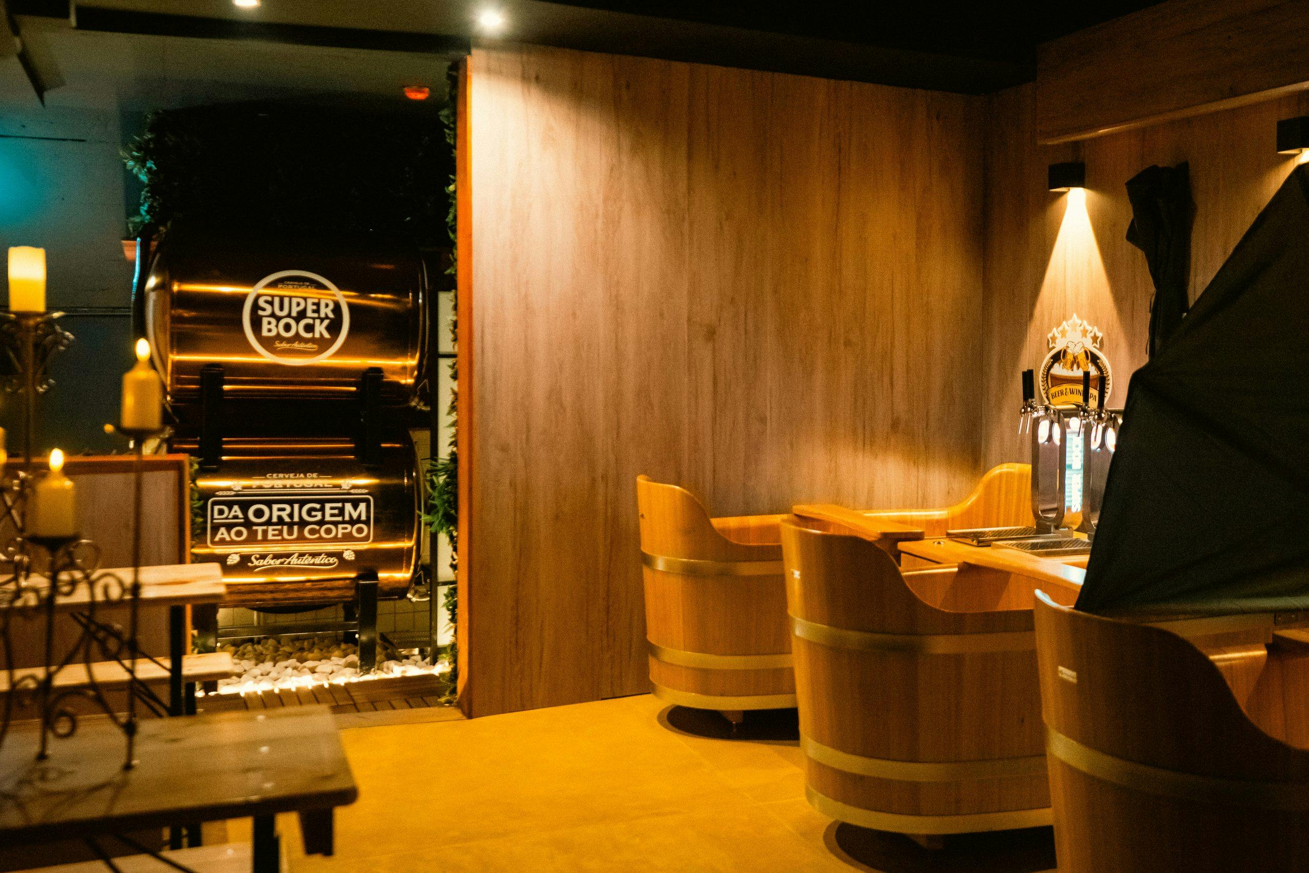 Dimly lit bar with wooden furniture, candle decor, a beer tap, and large beer barrels labeled "Super Bock."