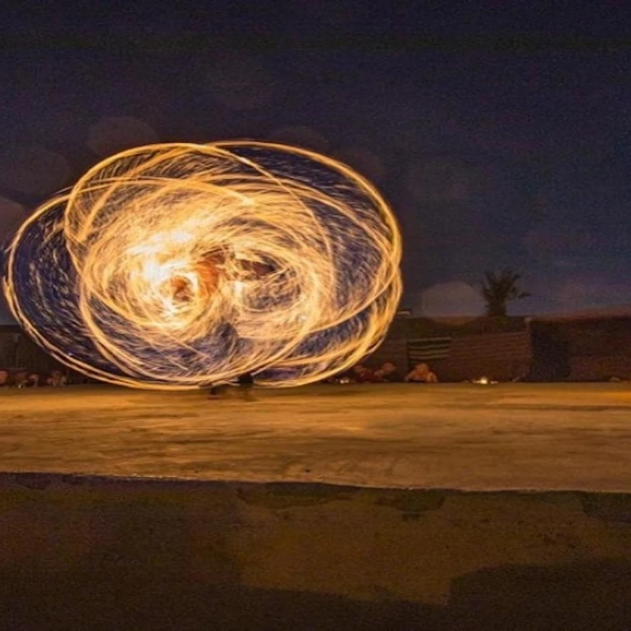 A performer spins multiple fire poi, creating swirling light patterns against a dark outdoor backdrop.